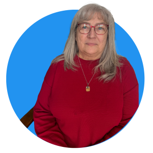 A woman with long gray hair and glasses is wearing a red sweater and a necklace, sitting in front of a blue circular background on a light gray backdrop.